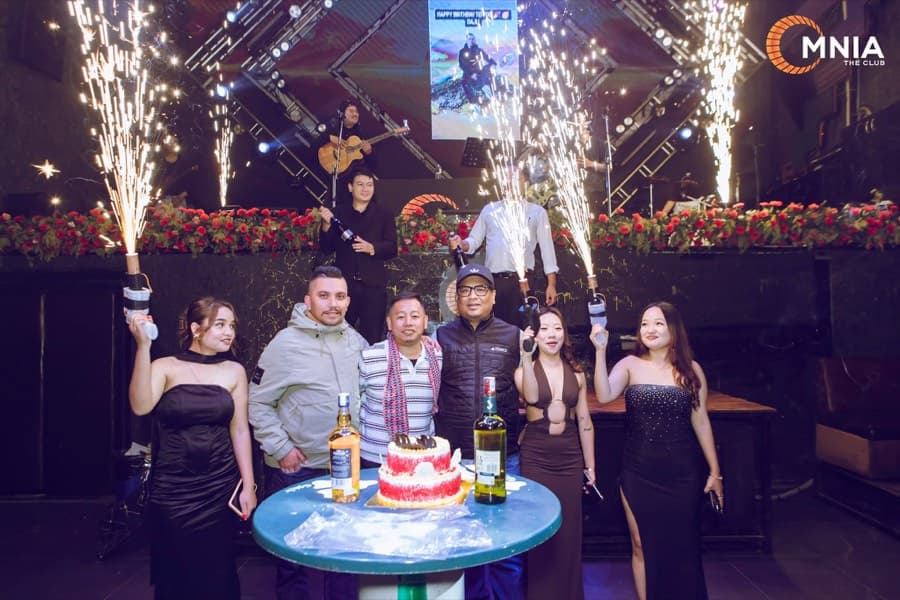 A birthday table — cake, bottles and six guests with fountains going off behind them