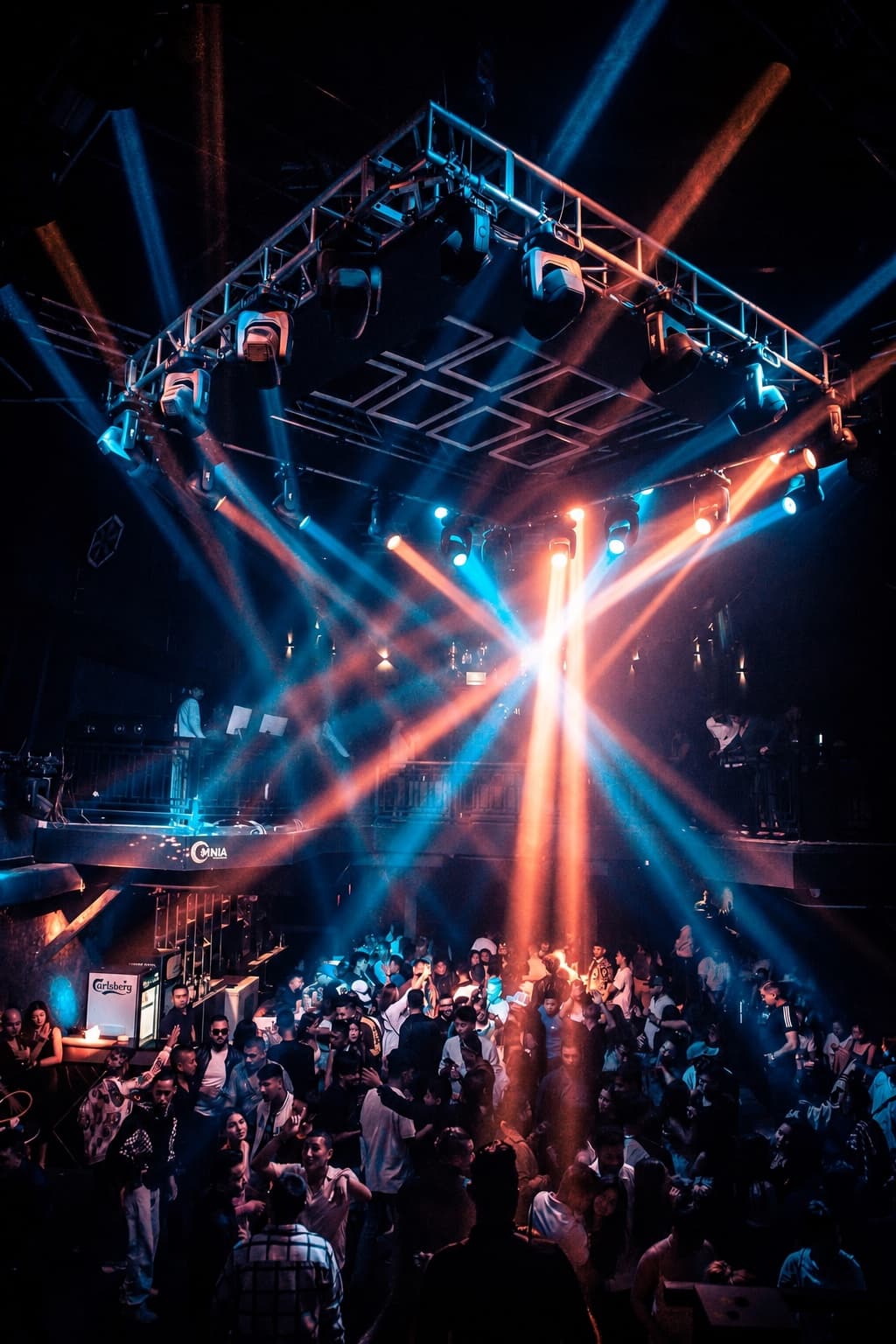 Blue and orange beams crossing over a packed OMNIA dancefloor, the mezzanine DJ booth and bar behind